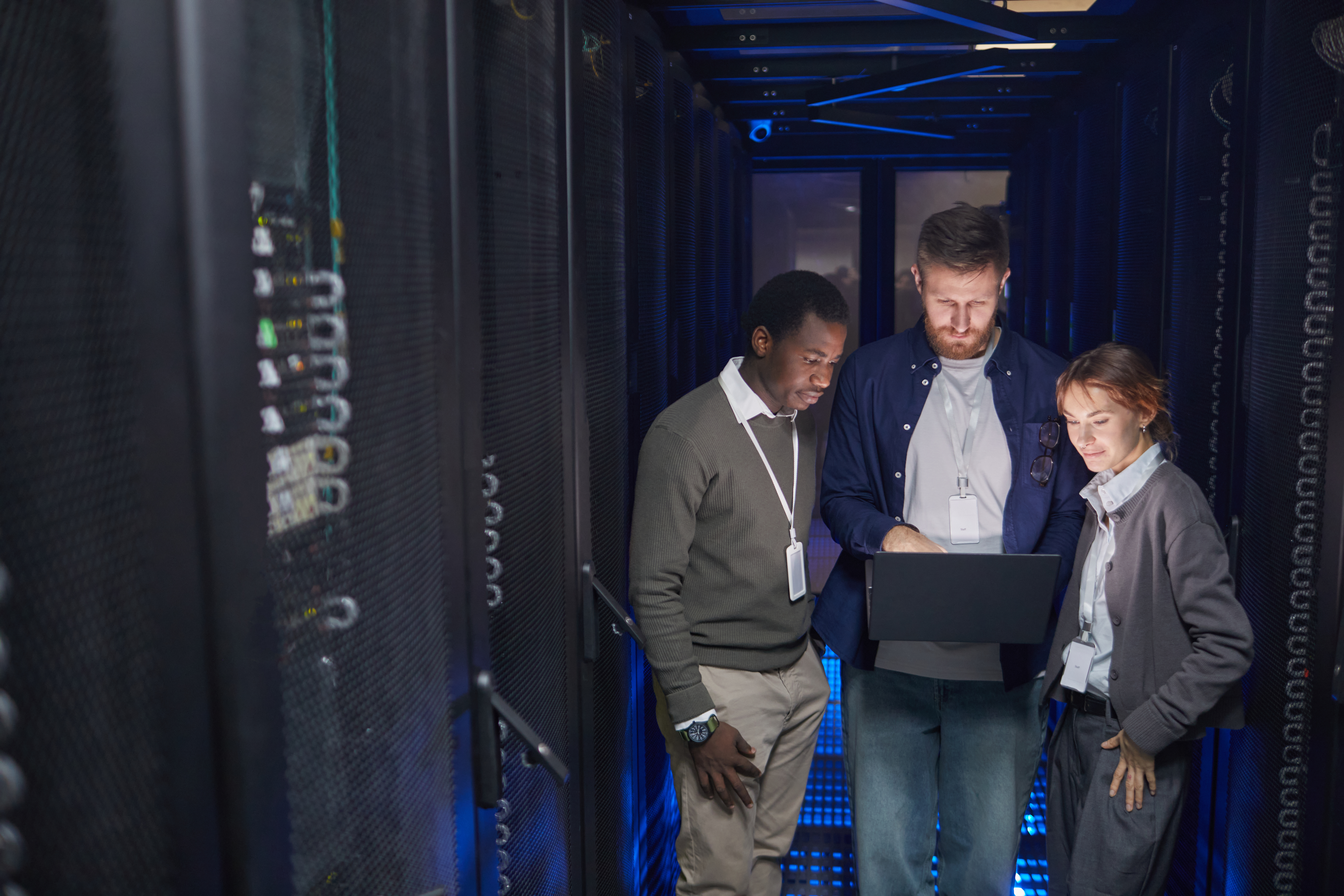 Group of diverse IT professionals standing in server room evaluating data on laptop while discussing network security and performance management issues